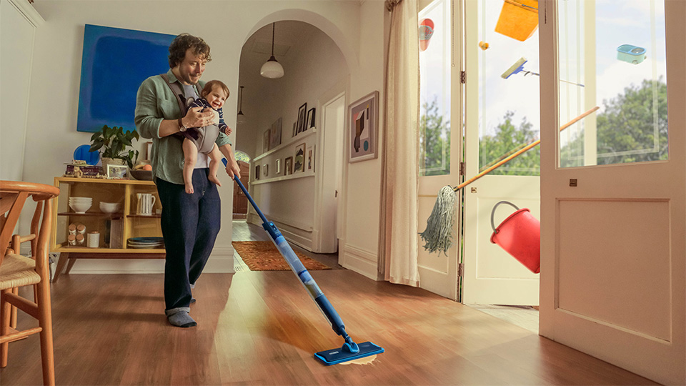 Ein Mann tr&auml;gt sein Baby und reinigt den Boden mit dem Philips OneUp elektrischen Wischmopp