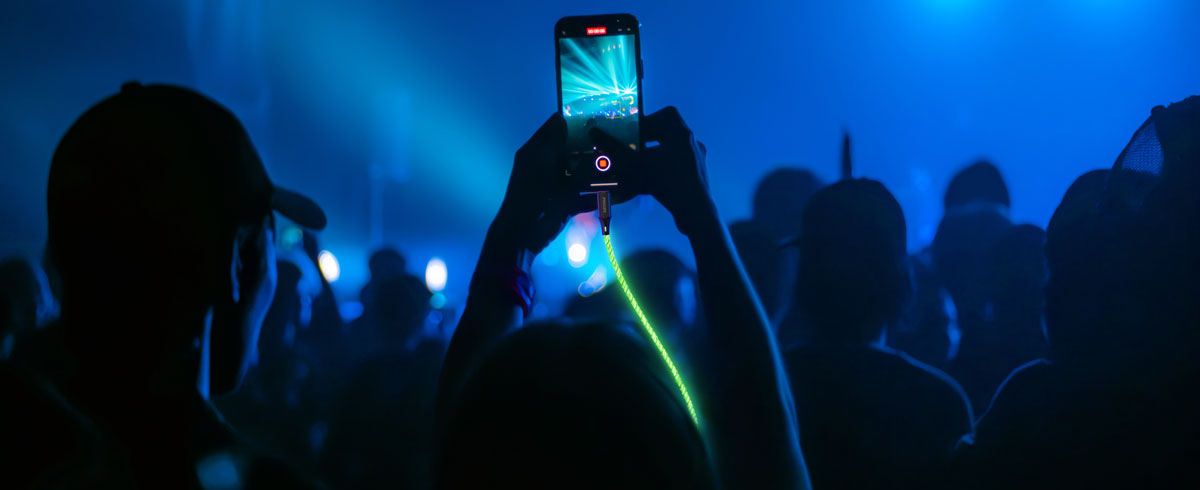 Eine Person h&auml;lt ein Handy, das mit einem leuchtenden Kabel verbunden ist, vor einer gro&szlig;en Menschenmenge und f&auml;ngt den Moment w&auml;hrend einer Veranstaltung ein.